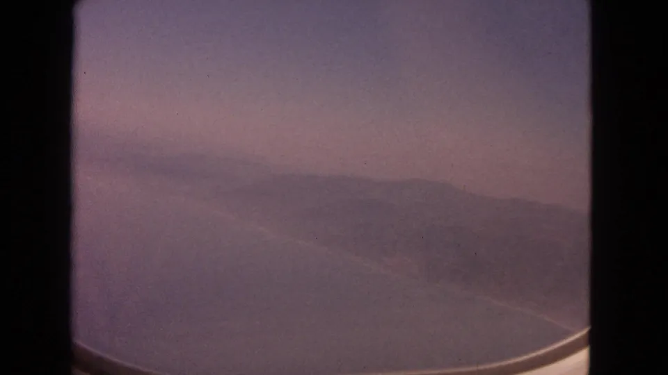 Vintage 8mm aerial view with muted sky, hazy hills, water below, and the curved edge of the airplane window.