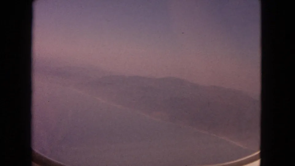 1963 8mm frame showing a hazy airplane-window view over water and low mountains under a pink-purple sky.