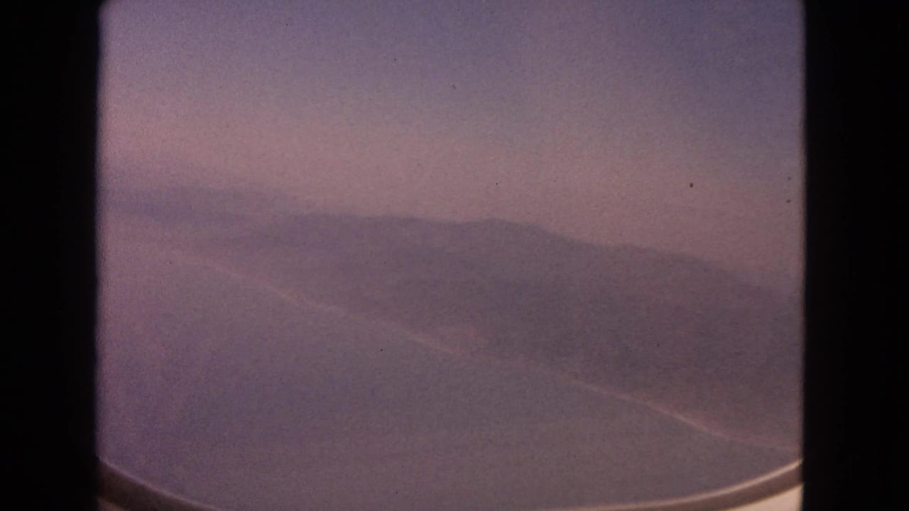 1963 8mm frame showing a soft, hazy view of ocean and distant land through an aircraft window.