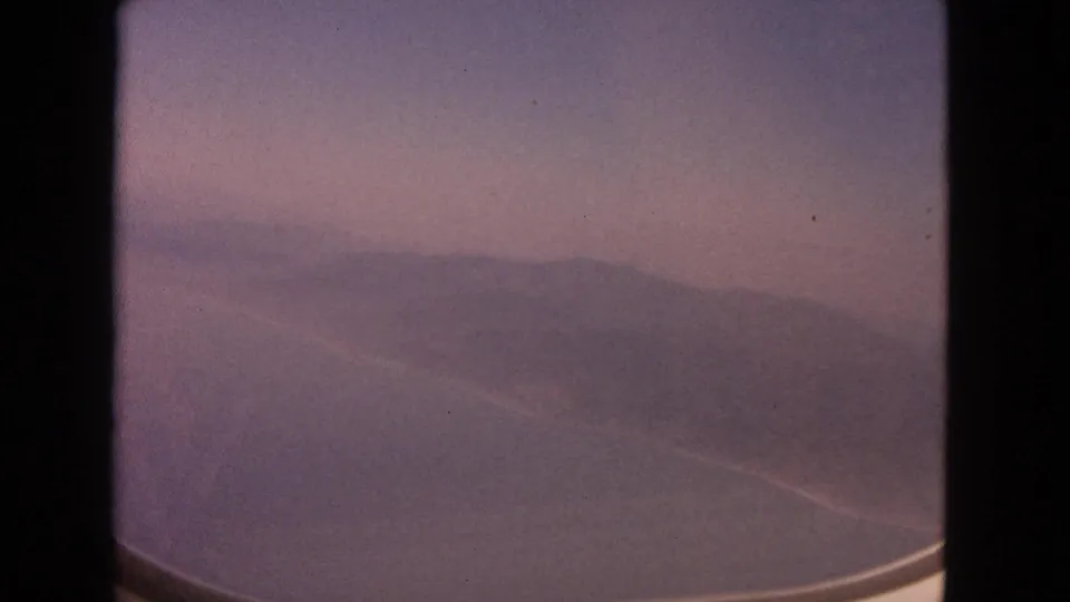 1963 8mm frame showing a soft, hazy view of ocean and distant land through an aircraft window.