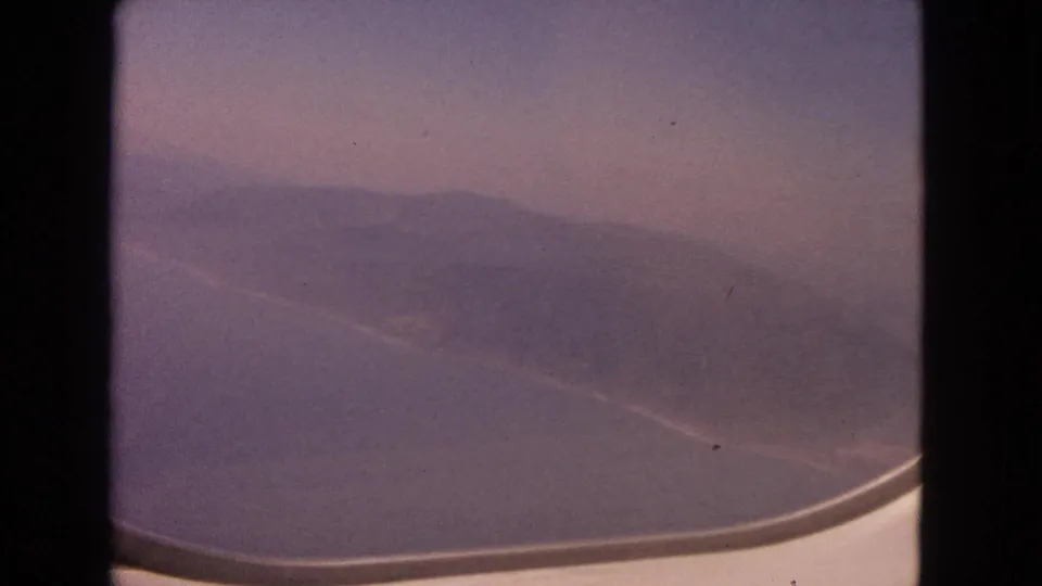 1963 8mm aerial view with a curved aircraft window edge framing hazy coastline and open water.