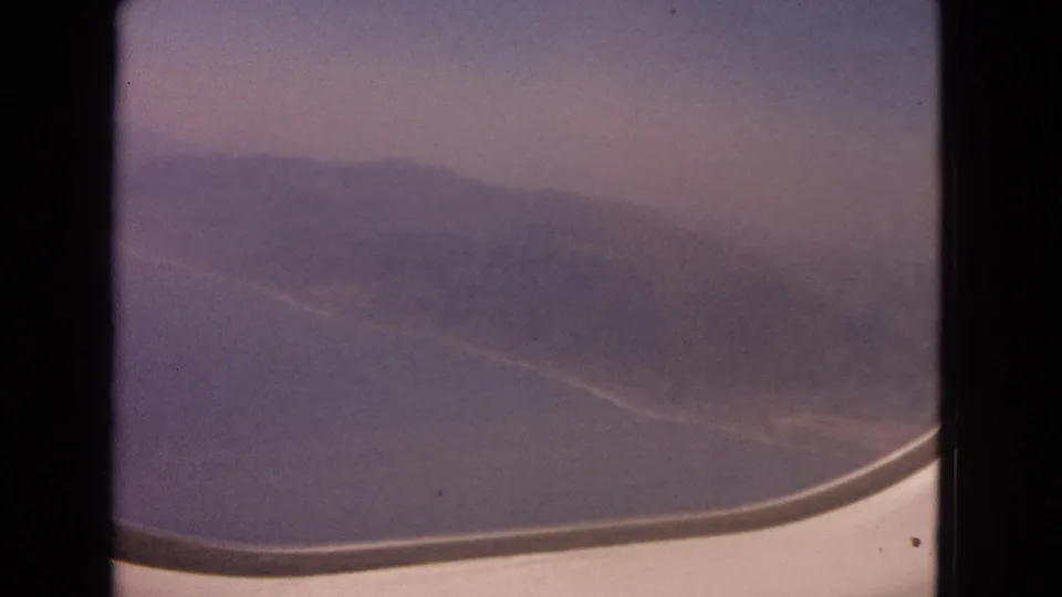 1963 8mm frame with a blurred shoreline, ocean surface, and curved airplane window in the foreground.
