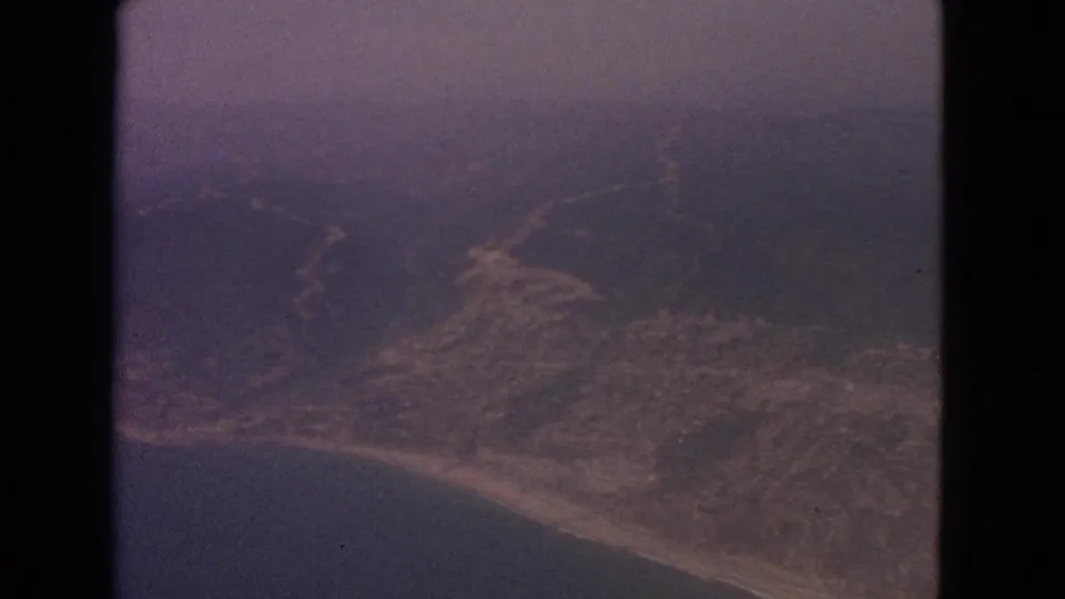 1963 8mm aerial frame showing coastal land, surrounding water, and a faint winding road or path below.