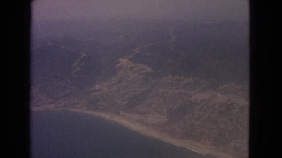 1963 8mm frame showing a hazy aerial view of shoreline, water, and inland terrain from an airplane-window perspective.