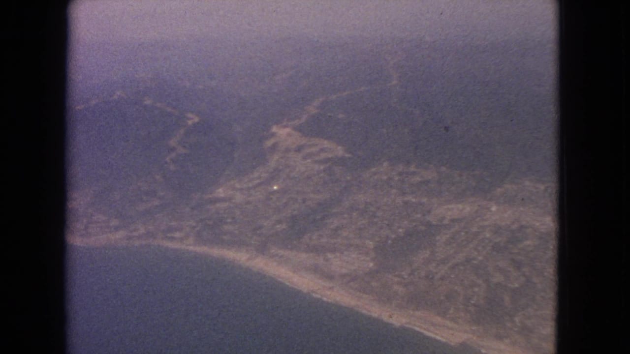 1963 8mm aerial frame with coastline, sea, inland hills, haze, and dark vertical film-strip edges.
