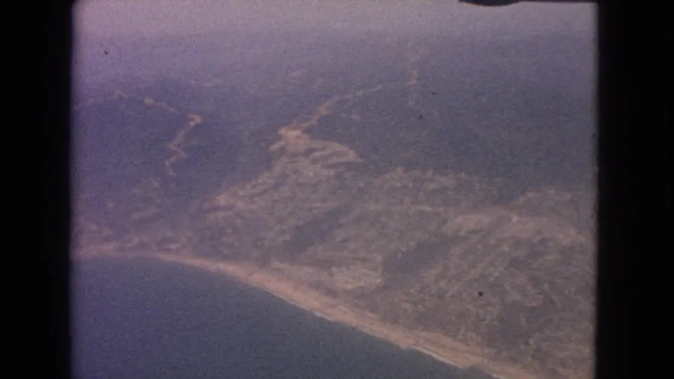 Grainy 1963 8mm frame showing a coastline and inland landscape from a high passenger viewpoint.