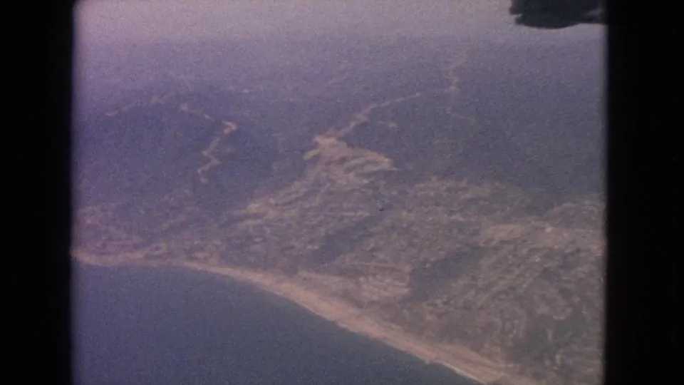 1963 8mm airplane-window view of a coastline, inland slopes, haze, and visible scanned film borders.