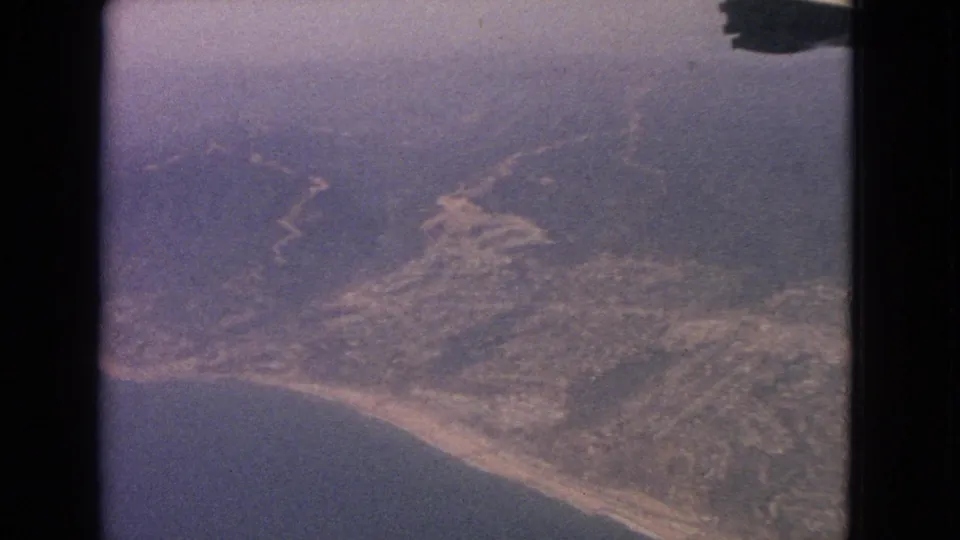 1960s 8mm airplane-window view over a hazy shoreline with hills, water, and scattered settlement below.