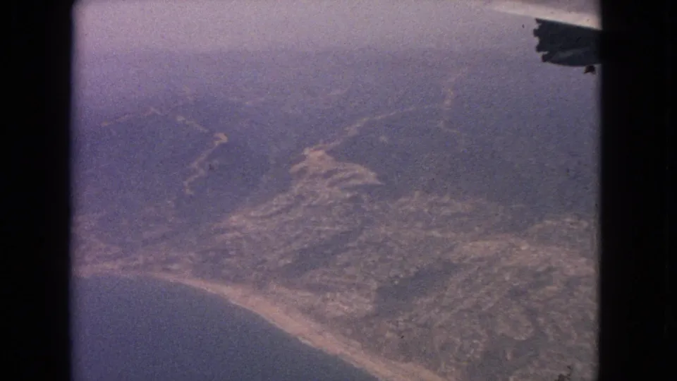 Vintage 8mm aerial frame showing a curving shoreline, muted water, and hill terrain from a moving airplane.