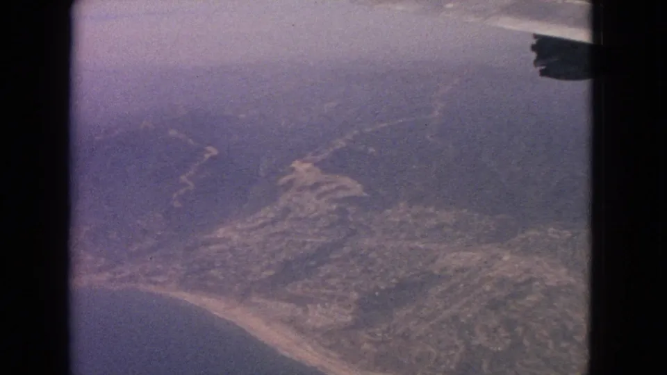 1960s home-movie aerial view over shoreline and hills, with muted color and a partial aircraft edge in frame.
