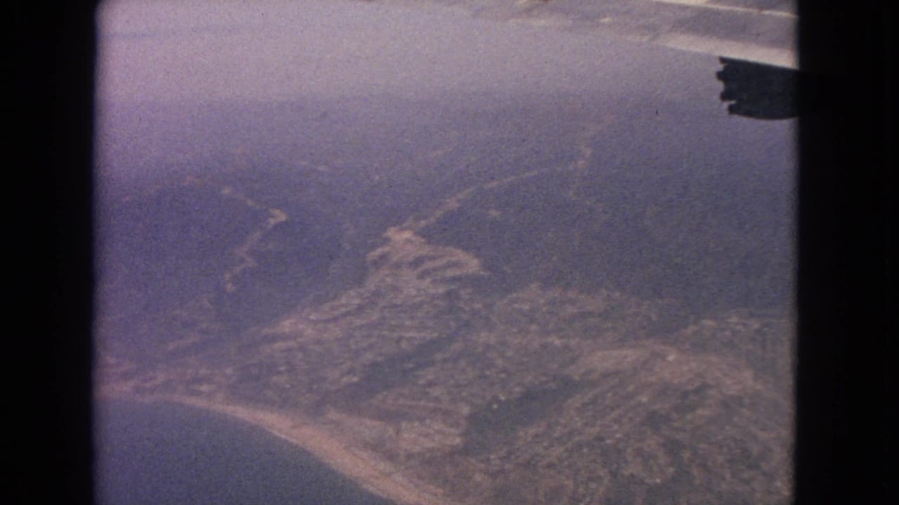 1963 8mm passenger-window view over a coastline with sandy beach, inland roads, and part of an aircraft wing visible.