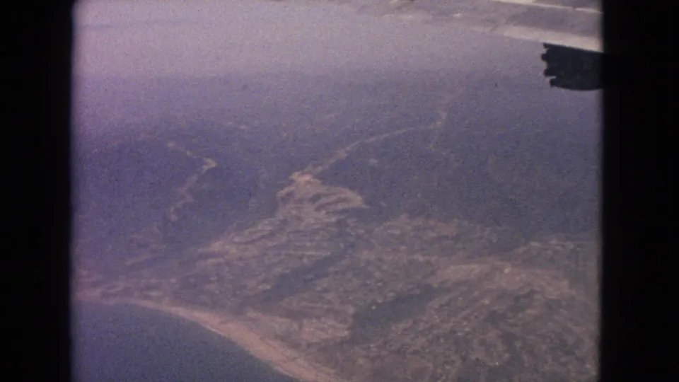 1963 8mm passenger-window view over a coastline with sandy beach, inland roads, and part of an aircraft wing visible.