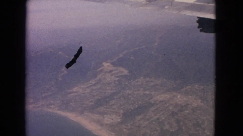 1963 aerial home-movie frame showing a coastal settlement, beach curve, ocean water, haze, and the aircraft wing edge.