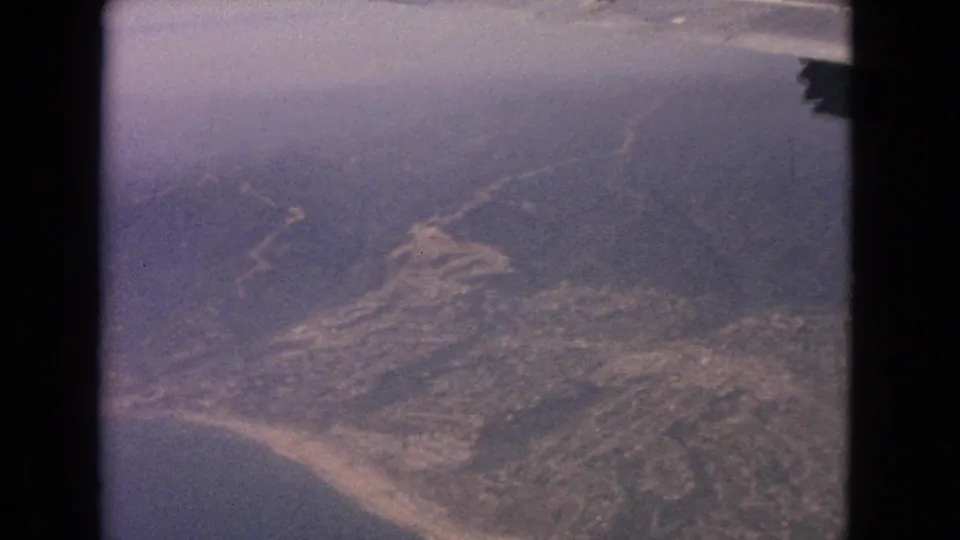 1963 8mm frame with a hazy airplane-window view of shoreline, ocean, inland roads, and developed terrain.