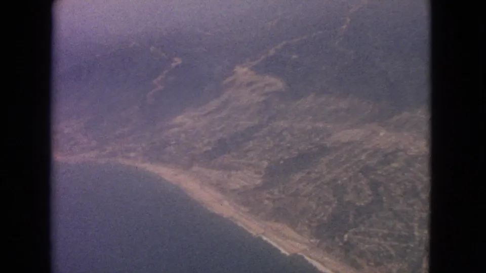 1963 8mm aerial view of a curved sandy shoreline, adjacent water, and inland development seen from a passenger aircraft.