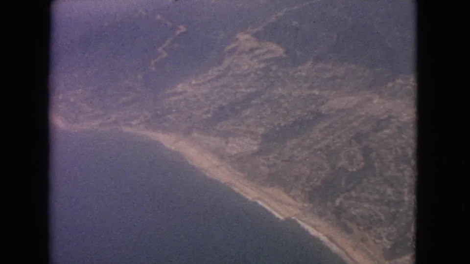 1963 8mm aerial view from an airplane over a rugged coastline with ocean, hills, haze, and window edges visible.