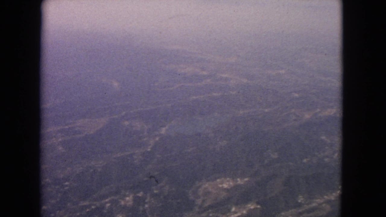 1963 8mm passenger-view aerial over hazy inland terrain with hills, faint roads or clearings, and dark window borders.