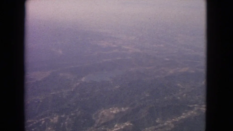 1963 8mm aerial frame with a faint coastline at left, broad inland hills, haze, film grain, and airplane-window framing.