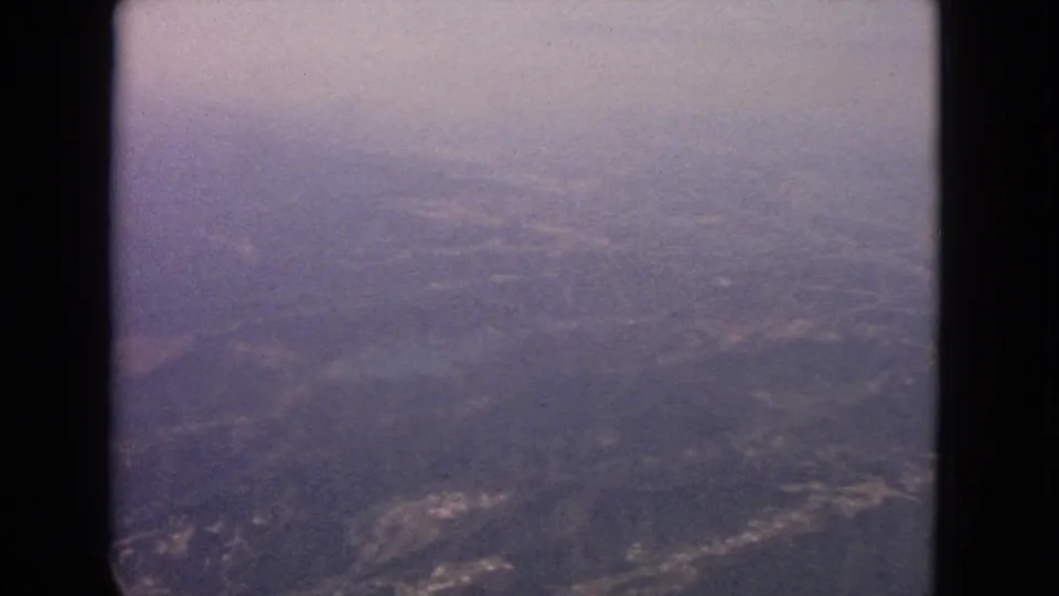 1963 8mm airplane-window view over a distant hazy landscape with rolling terrain, pale clearings, and soft blue-purple color.