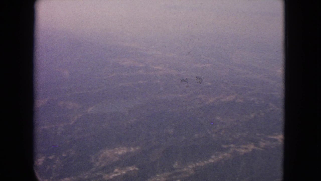 1963 8mm view from an airplane window over hazy terrain near Barrington, Illinois, with the aircraft window edge framing the scene.