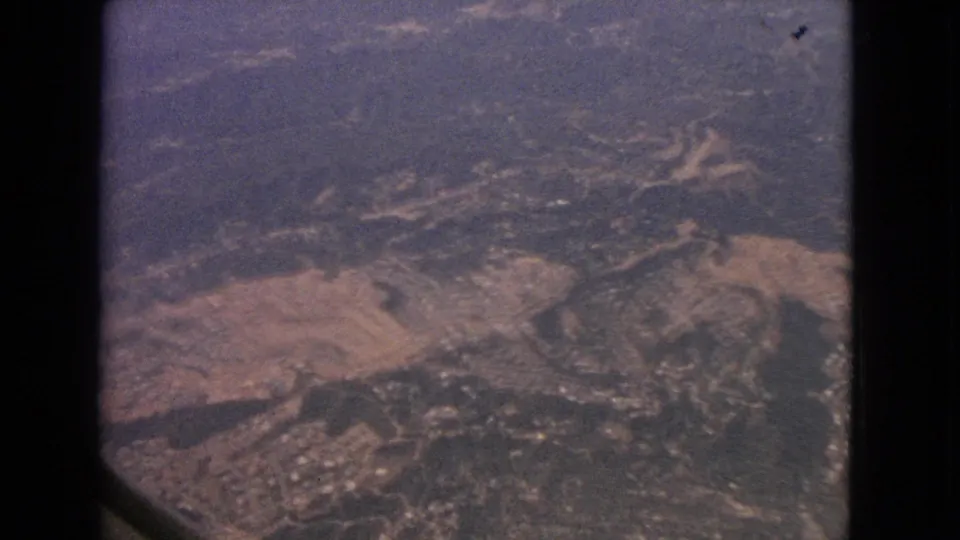 1963 8mm aerial view through the aircraft window, showing fields, roads, wooded areas, and clustered development below.