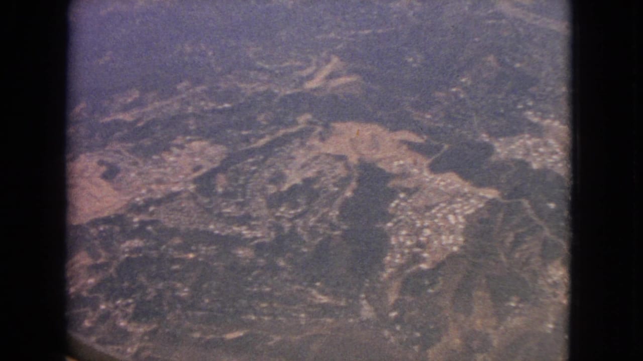 1963 8mm aerial frame showing rugged terrain, winding roads, pale clearings, and small settlement clusters from an aircraft window.