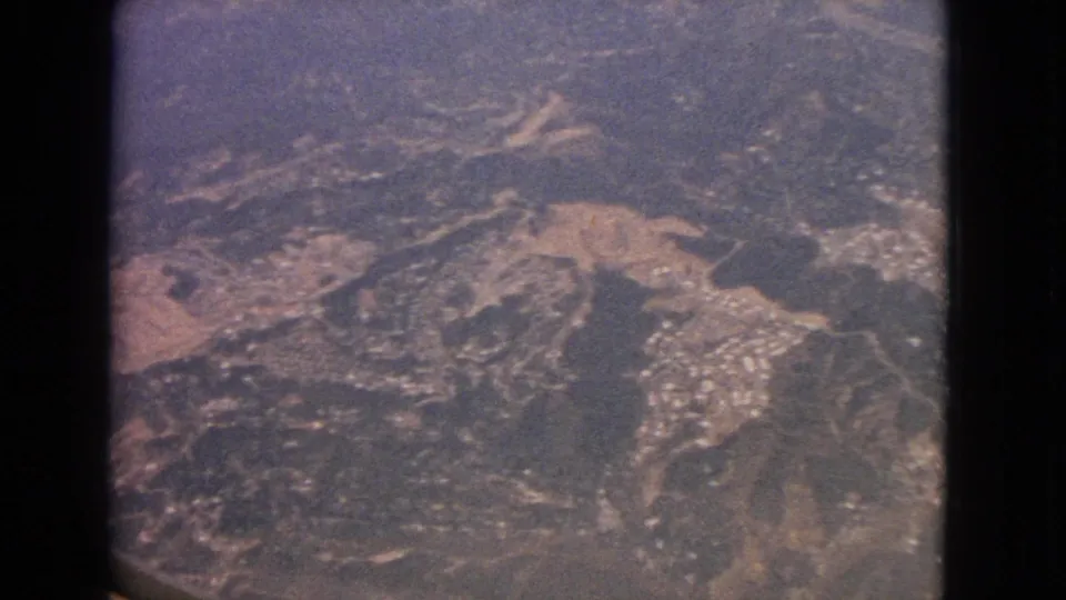 1963 8mm aerial frame showing rugged terrain, winding roads, pale clearings, and small settlement clusters from an aircraft window.