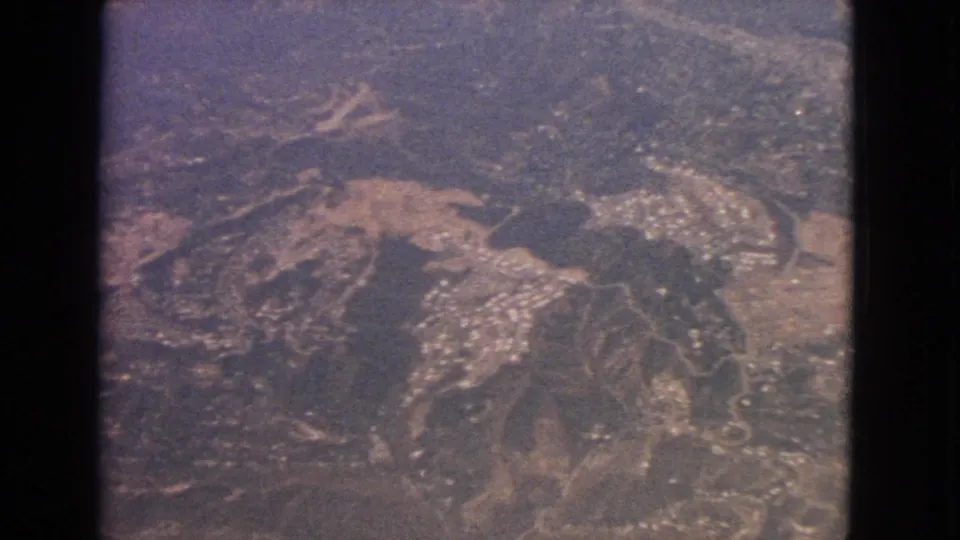 1963 8mm aerial view of a compact settlement among hills, with roads and cleared ground visible below.
