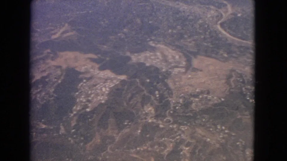 1963 8mm frame looking down on rugged slopes, road curves, open land, and scattered structures from an aircraft.