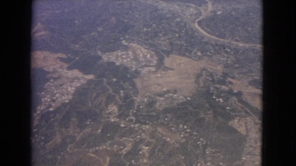 1963 8mm aerial frame showing rugged hills, pale road lines, open patches, and distant settlement clusters below.