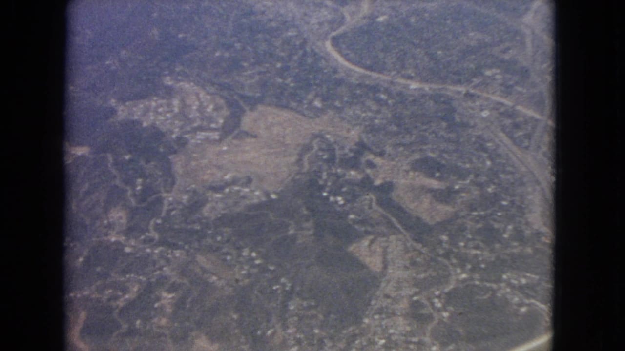 Aerial landscape with curved roads, open land, woods, and scattered structures.