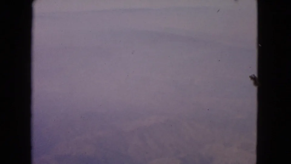 1963 8mm home-movie frame showing a hazy airplane window view over clouds, with dark window framing and soft purple color.