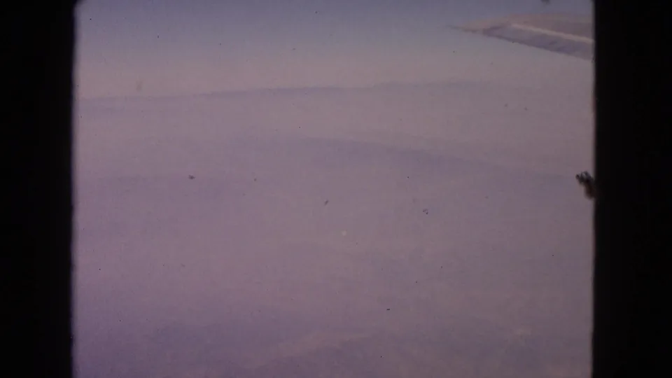 1963 8mm frame from an airplane window, showing soft cloud cover and a partial wing at the upper right.