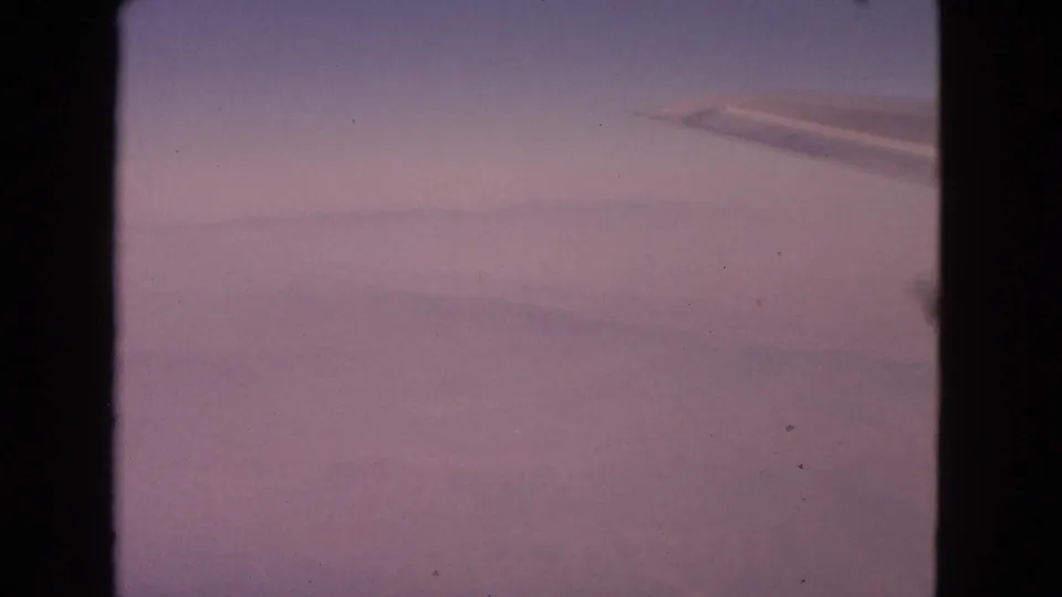 1963 8mm home-movie view from a passenger window, with wing detail, cloud cover, and dark window framing.