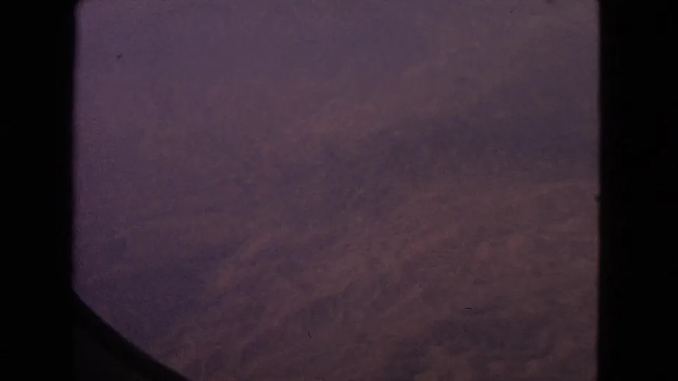 1963 8mm frame showing a darker aerial view through an airplane window, with hazy ground or cloud texture below.