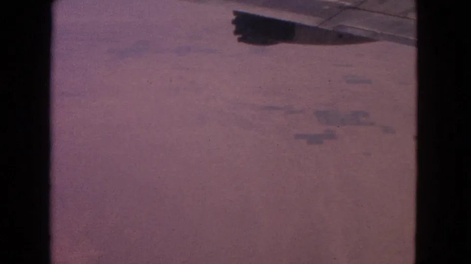Vintage 8mm frame showing a passenger-flight wing edge and reddish aerial terrain, with visible grain and color shift.