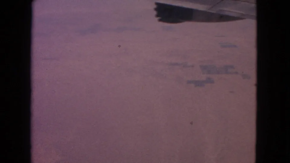 1963 home-movie airplane-window frame with wing silhouette, reddish terrain, and soft analog 8mm texture.