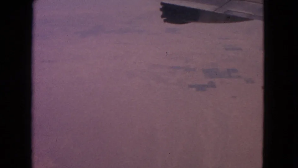 1963 8mm passenger-window view from an airplane, with the wing edge over a hazy purple aerial landscape.