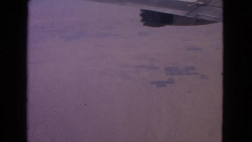 8mm aerial passenger view with a wing edge and faint ground divisions under a purple-tinted haze.