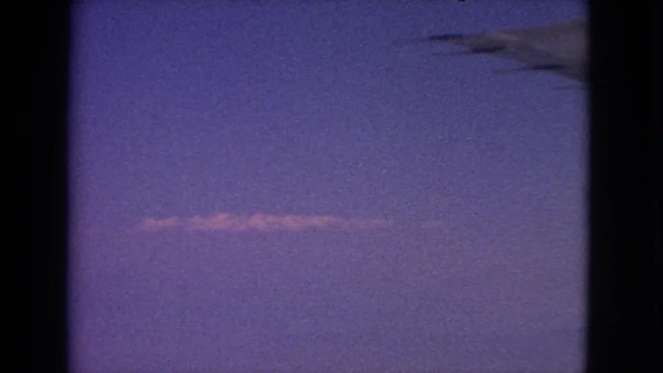 1963 8mm sky view from an airplane window, showing a dark wing shape and a thin contrail below.