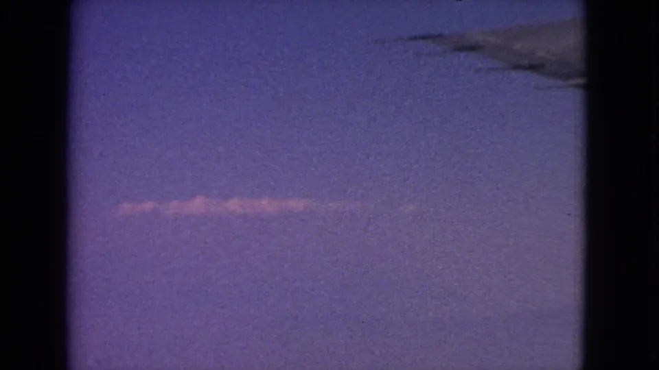 1963 8mm frame of a pale contrail below a partial airplane wing, seen through an aircraft window.