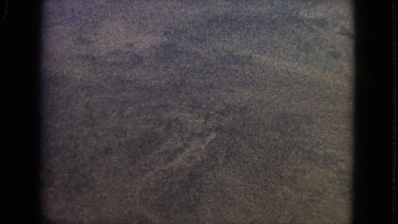 Dark grainy airplane-window view with indistinct ground or cloud texture.