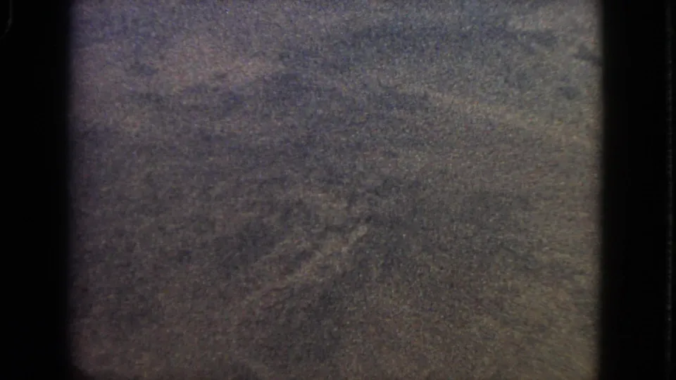 1963 8mm frame from an airplane window, showing a dark, grainy, indistinct aerial view with window-edge shadows.
