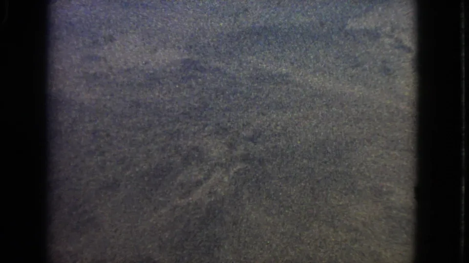 1963 8mm airplane-window view with dark framing and indistinct aerial terrain or cloud texture below.