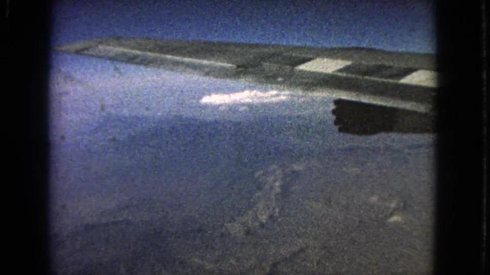 1963 8mm passenger-window view showing an airplane wing against blue sky with a bright cloud below.