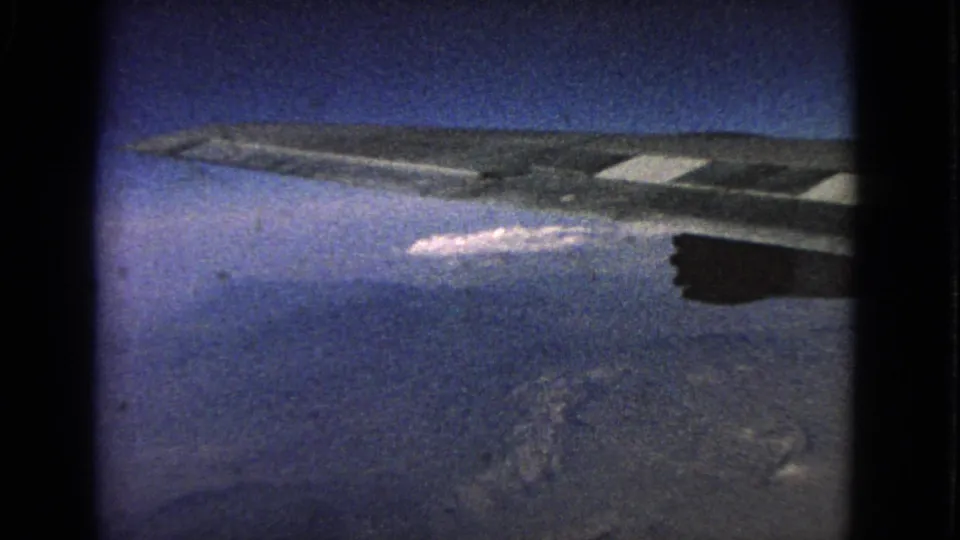 1963 8mm passenger-window frame with an airplane wing, dark engine shape, blue sky, and distant cloud below.