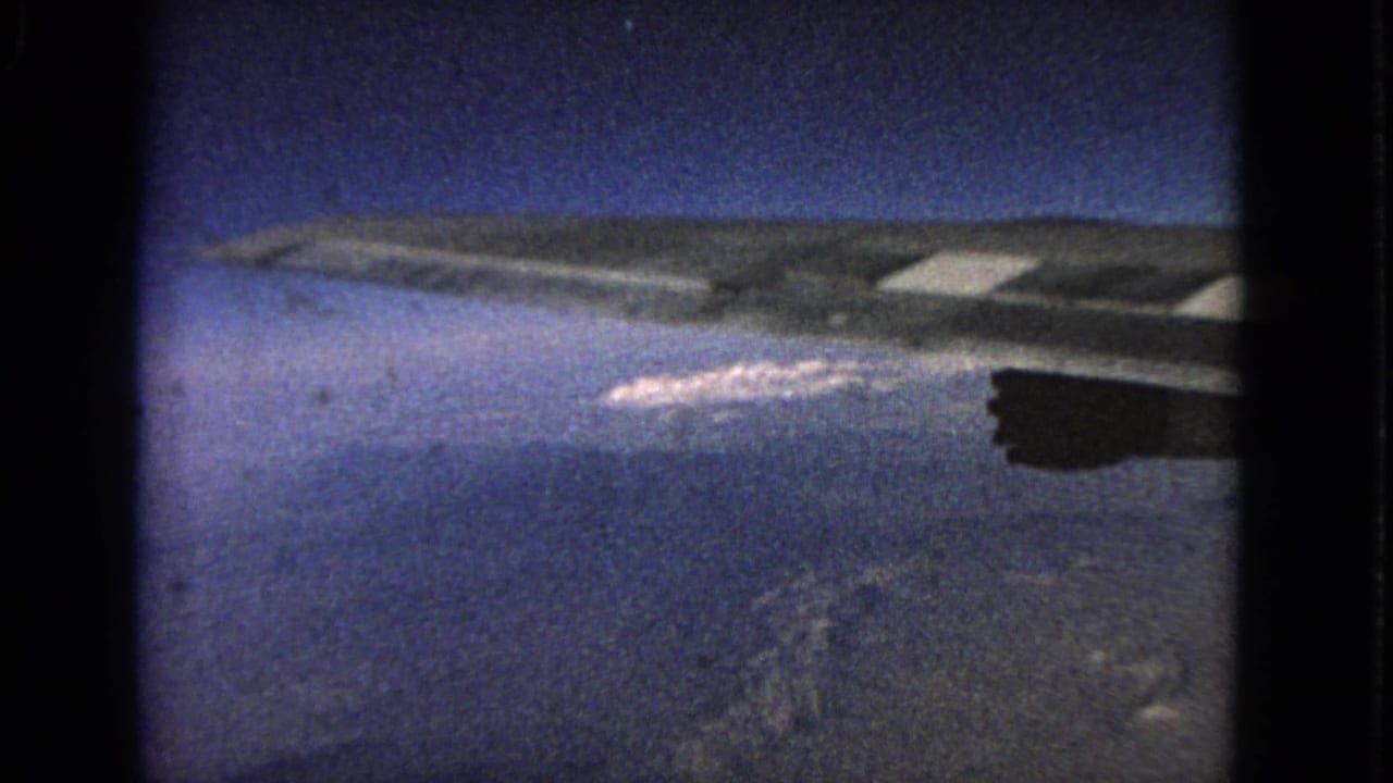 Aircraft wing and engine seen above clouds and distant terrain through an airplane window.
