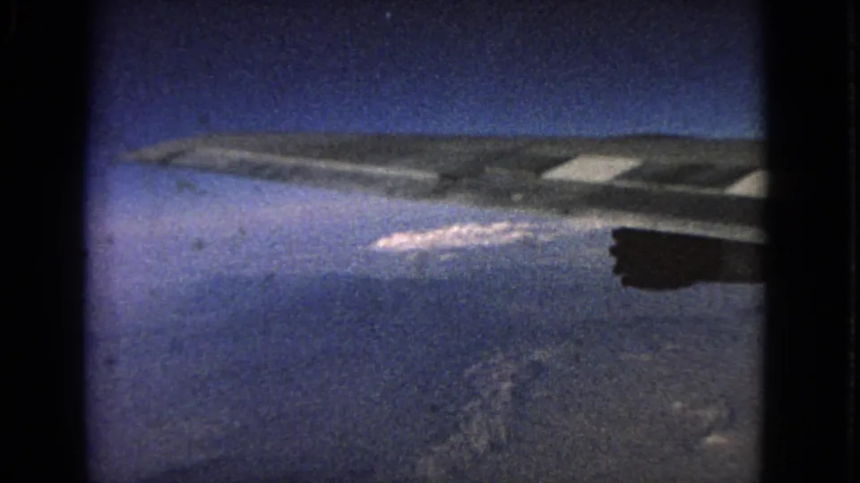 1963 8mm passenger view from an airplane window, showing the wing, engine, blue sky, and distant clouded landscape below.