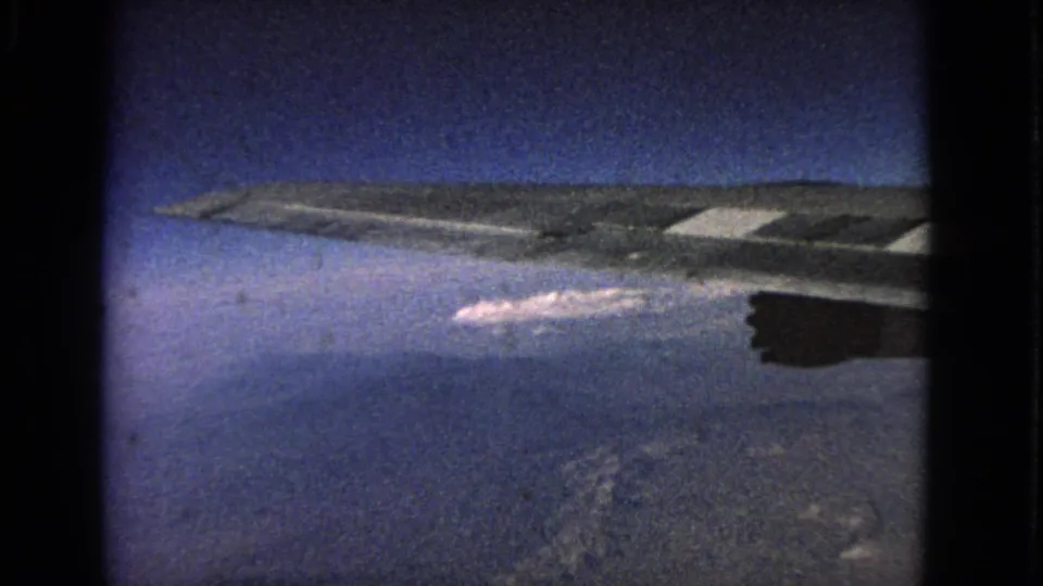 1963 8mm in-flight window view with an aircraft wing crossing the frame above clouds and a distant landscape or water surface.