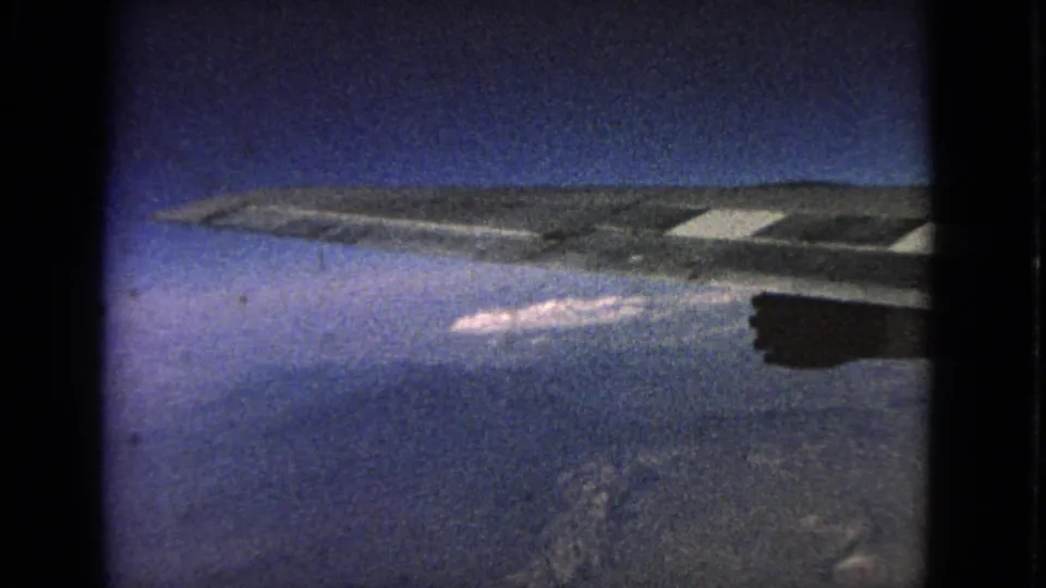 1963 8mm airplane-window frame showing the wing and engine silhouette over a hazy cloud layer and distant surface below.